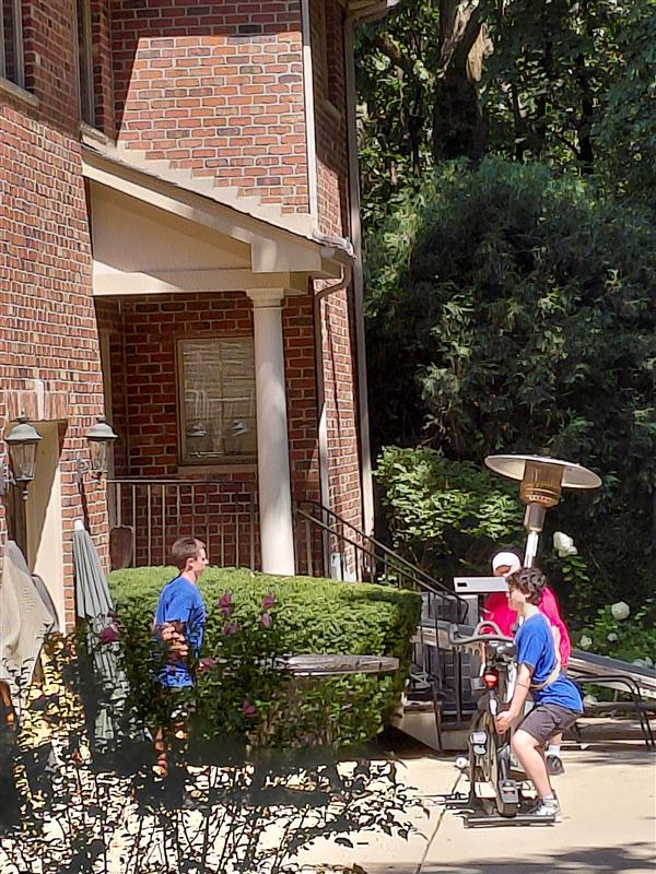Two children engage in outdoor activities, one exercising on a stationary bike while another talks nearby. A brick house and greenery provide context.