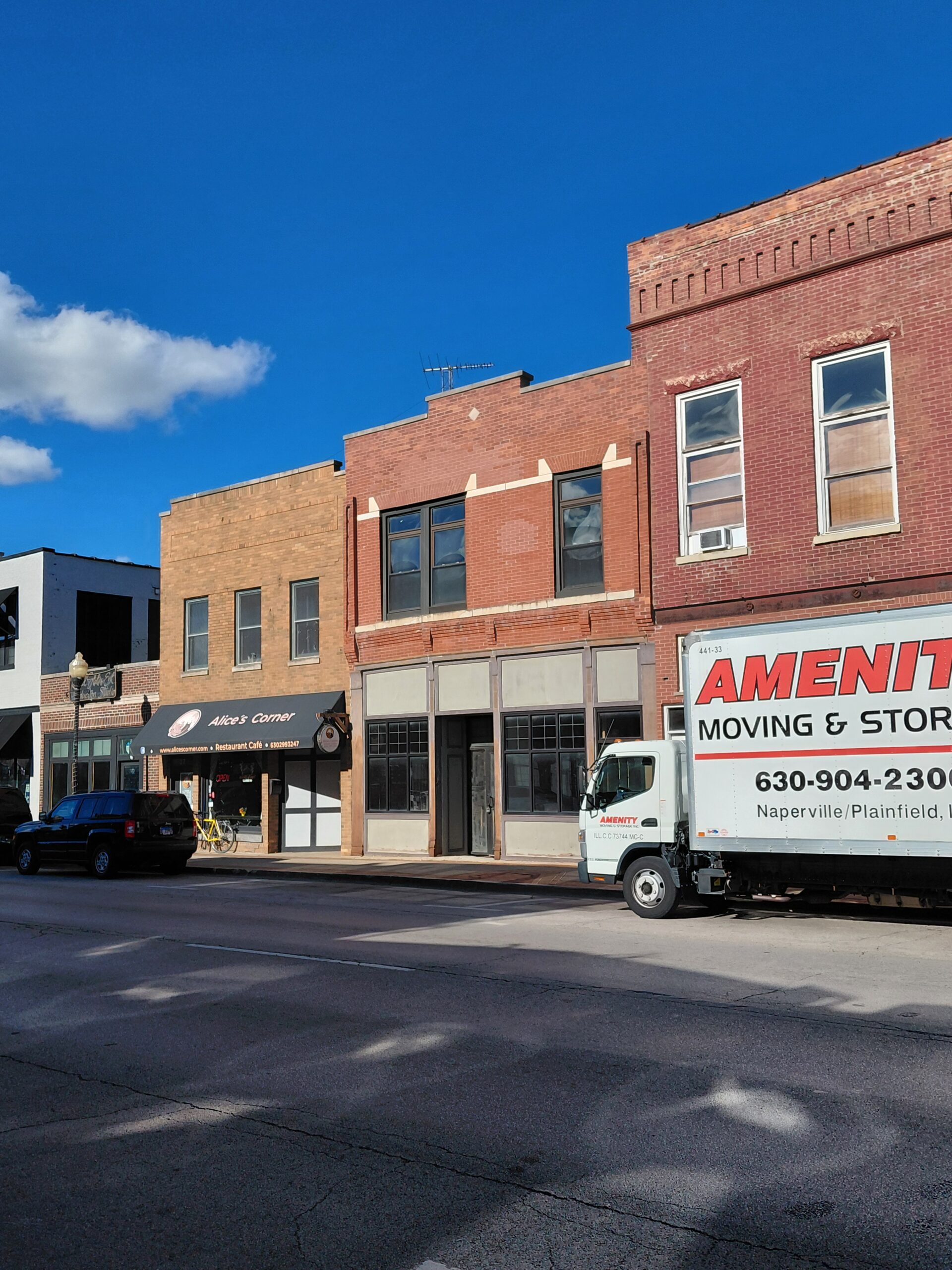 Historic storefronts line a street under a blue sky, featuring "Al's Corner" restaurant and a moving truck parked nearby.