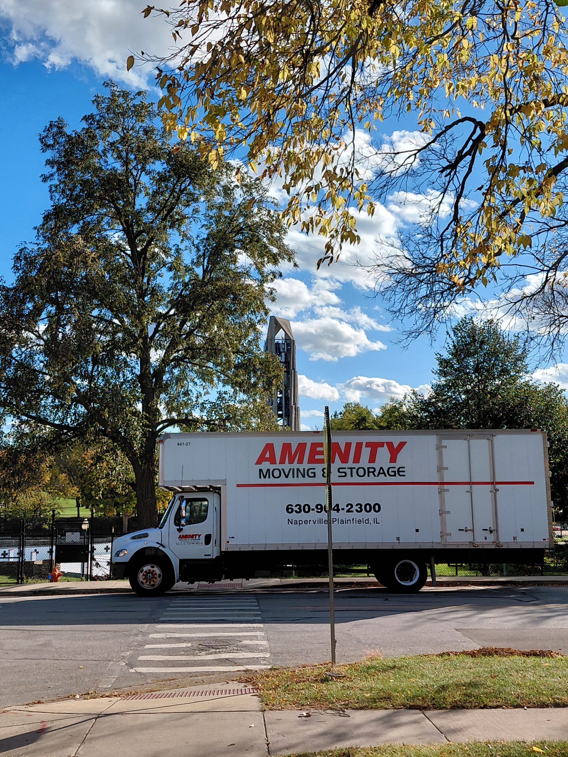 Moving truck labeled 'Amenity Moving & Storage' parked on a street, surrounded by autumn foliage and a clear blue sky, indicating its service in the area.