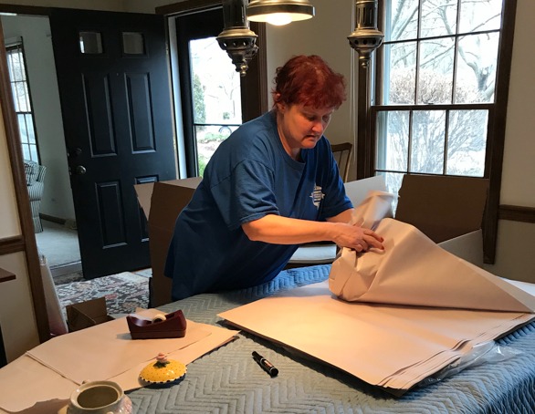 A woman in a blue shirt carefully folds packing material on a table, surrounded by boxes, preparing items for moving. Natural light streams in through large windows.
