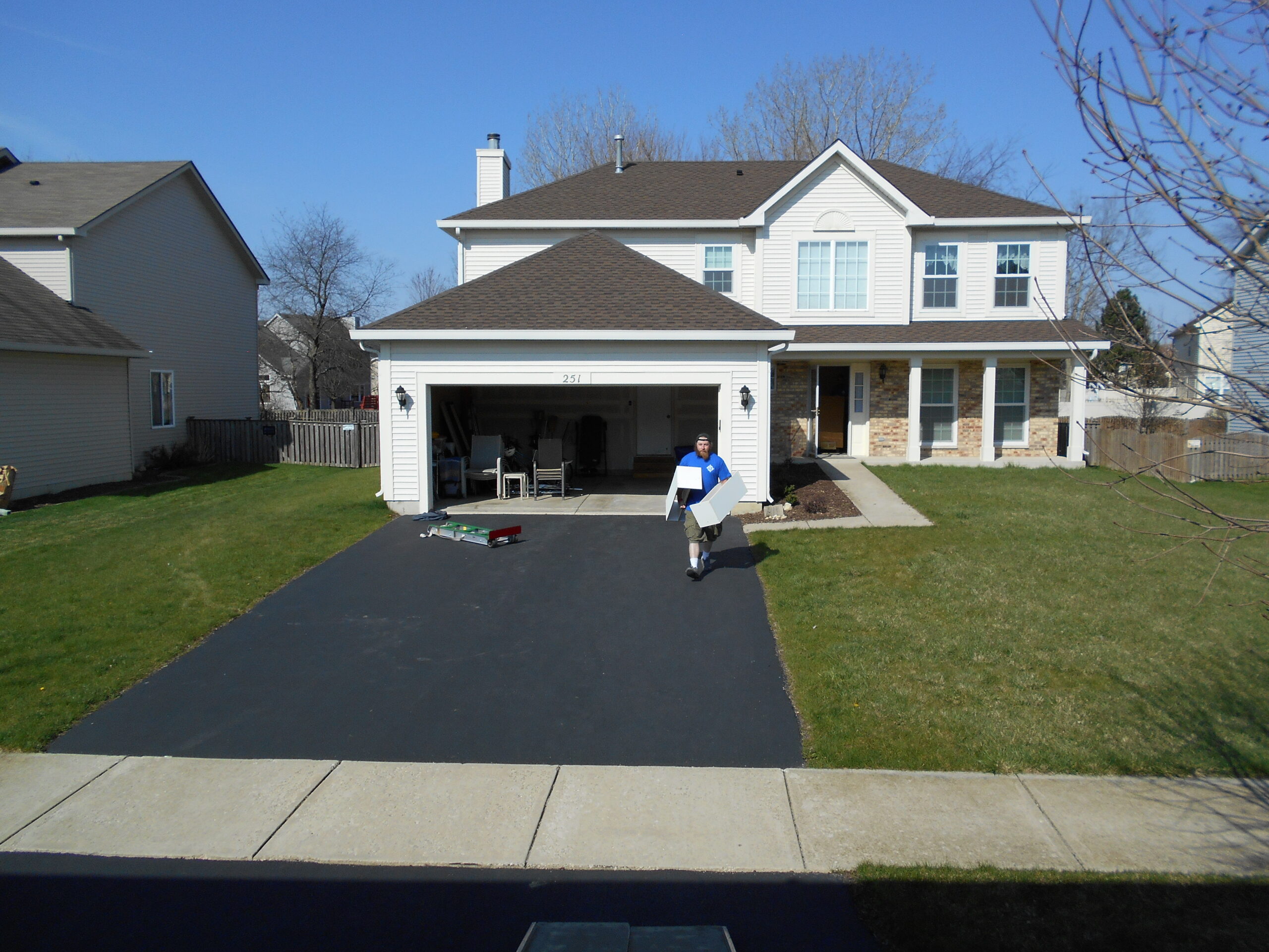A person in a blue shirt carries a large white item in front of a two-story suburban house on a sunny day.