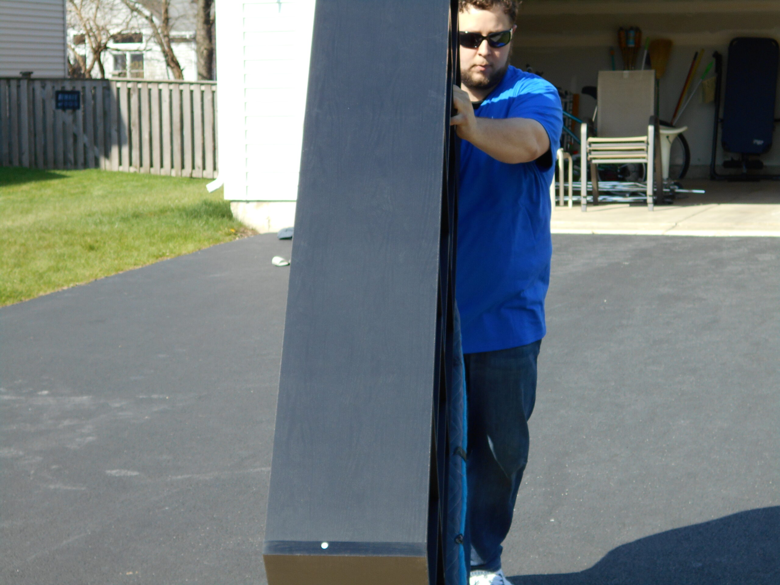 A man in a blue shirt and sunglasses carefully maneuvers a large object in a driveway, showcasing the effort of moving heavy items.