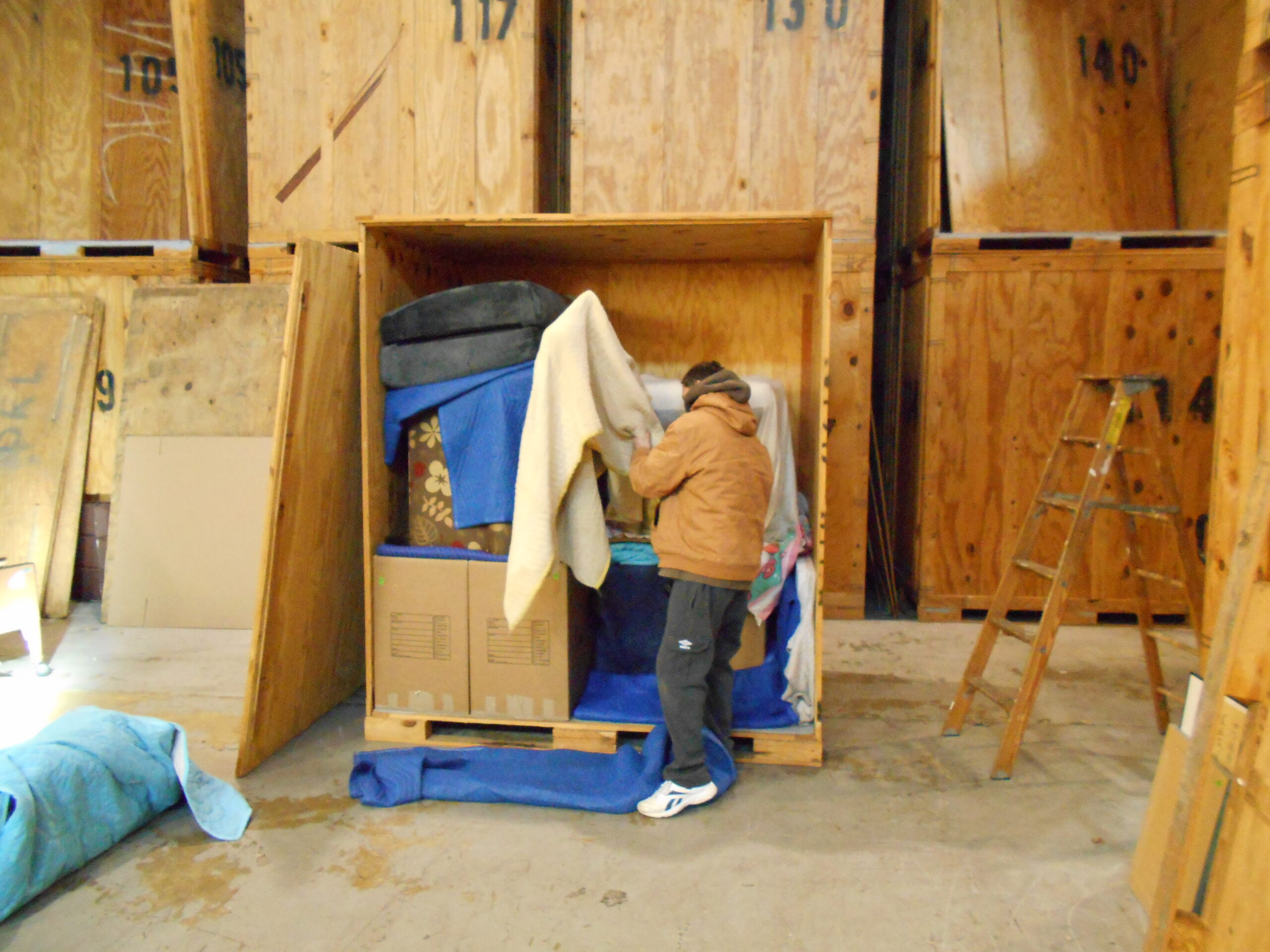 A person in a brown jacket organizes items inside a wooden storage unit filled with blankets and boxes. A ladder is visible nearby.