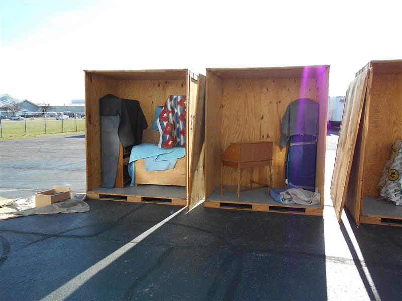Three wooden storage units display various items: a garment, colorful blankets, and a small wooden table, set in a parking area.
